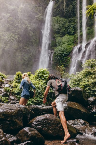 Couple crossing a stream together in the forest on a hike. Hiking man and man crossing a river. Climbing big rocks.